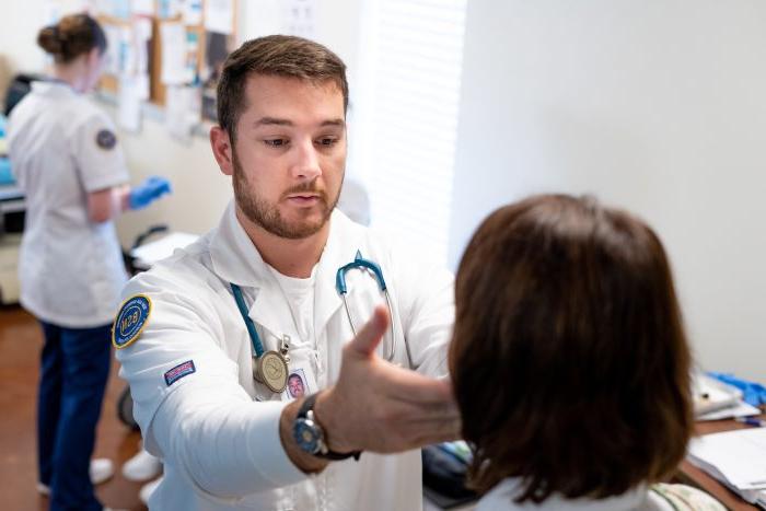 Student in white lab coat reaches out hand to patient's face during checkup.